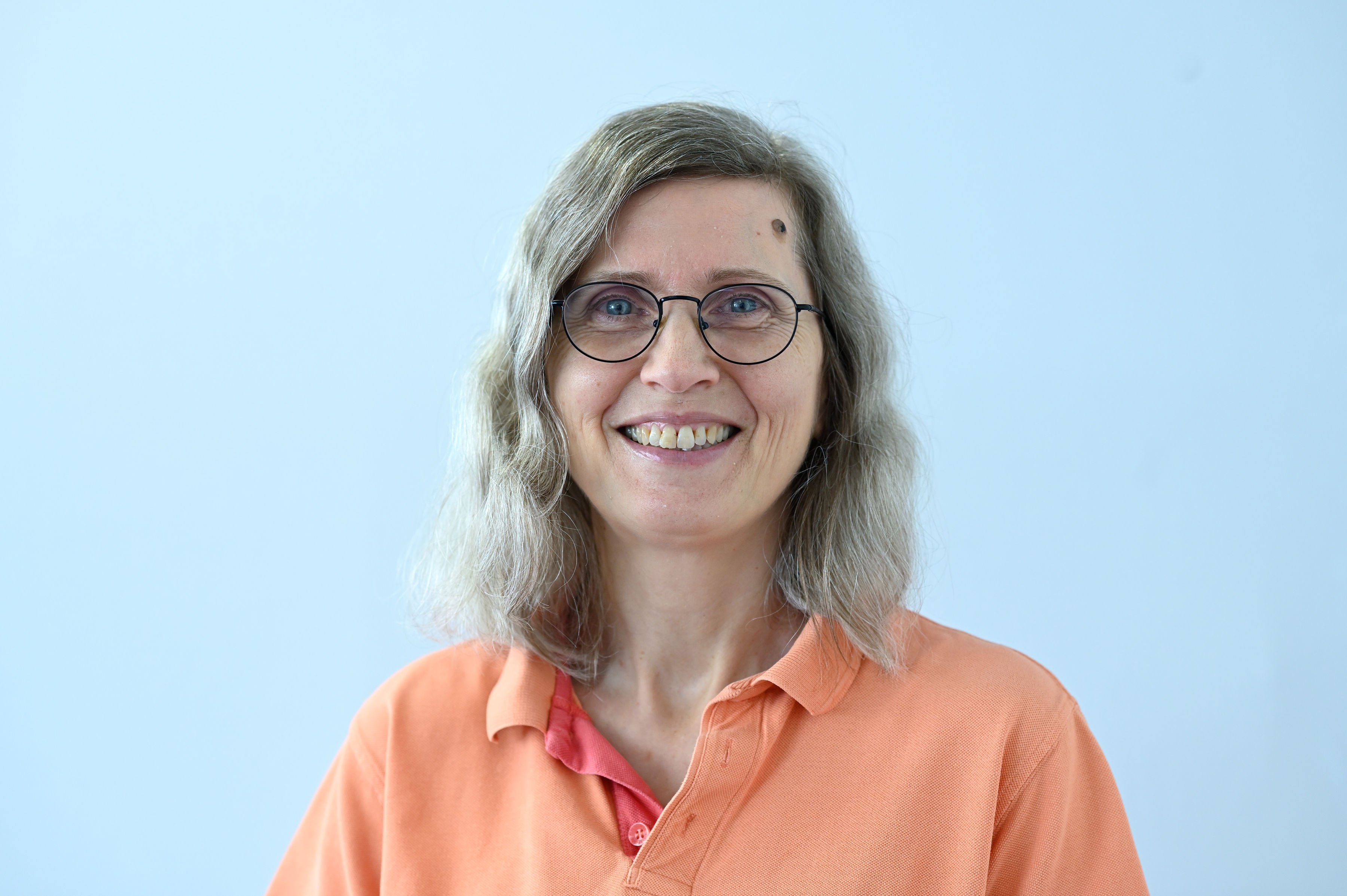 a lady in her 50s with Shoulder length brown and grey hair, an open smile and glasses.